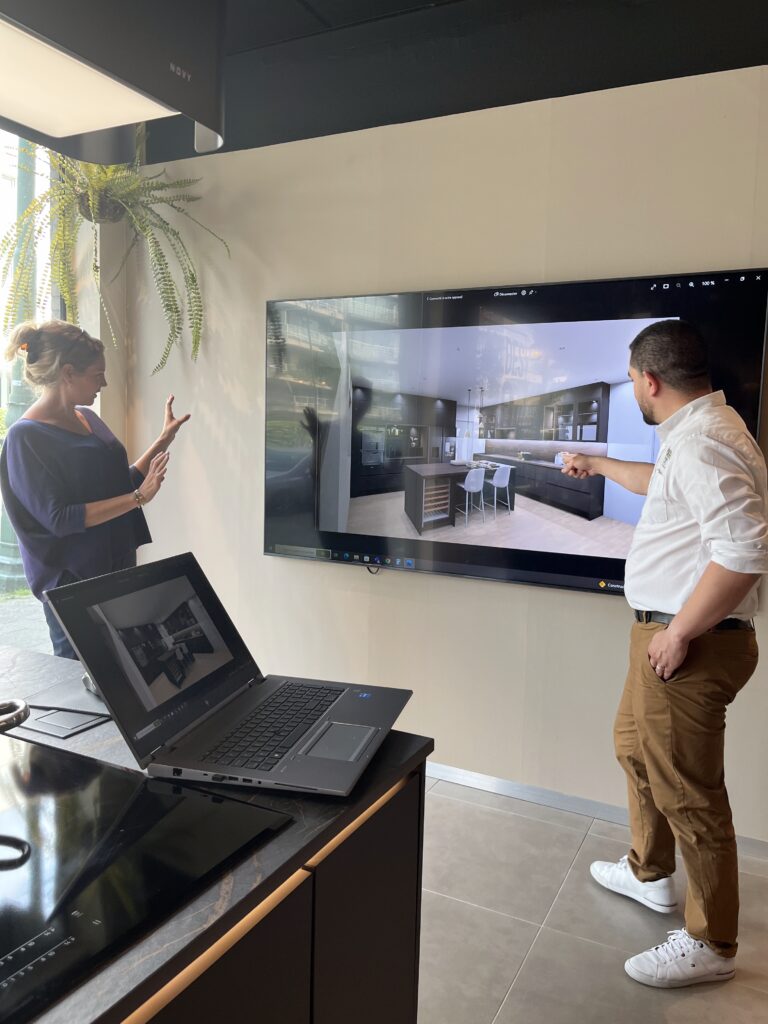 Deux personnes discutent devant un écran affichant une cuisine moderne, avec un ordinateur portable ouvert sur un plan de travail sombre.