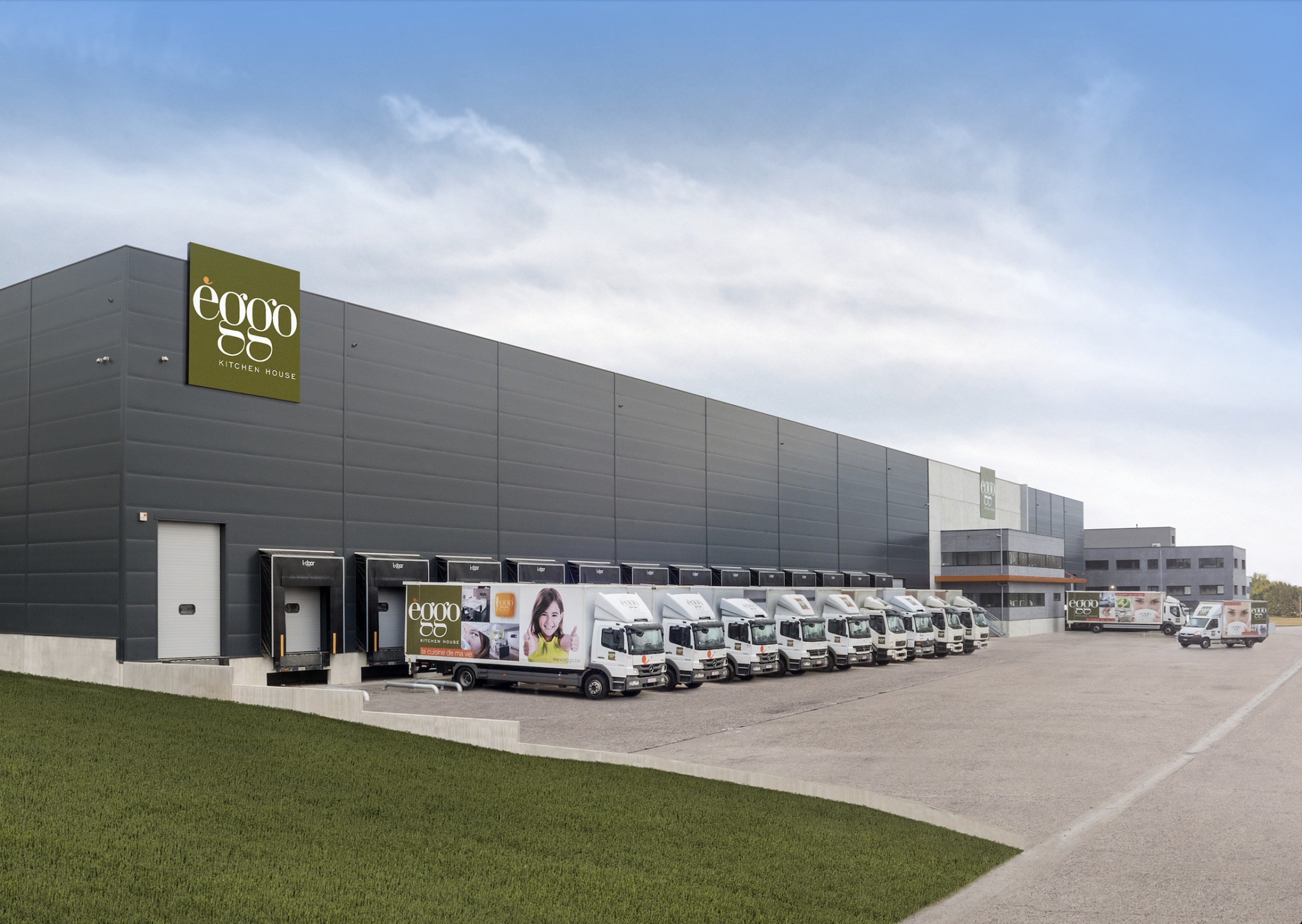 Un bâtiment industriel gris avec des camions blancs alignés devant des quais de chargement, ciel partiellement nuageux.