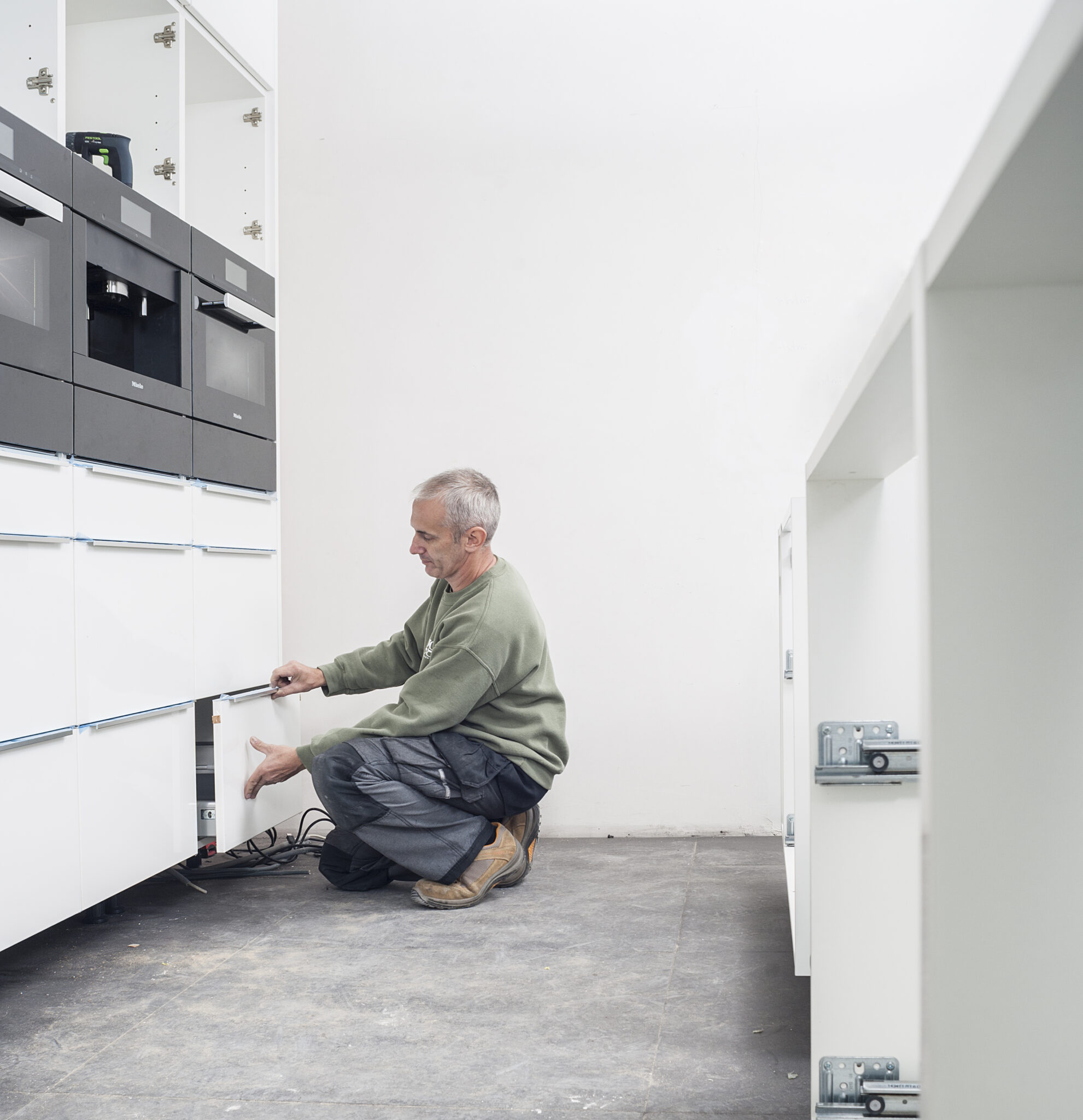 Un homme en pull vert installe des tiroirs blancs dans une cuisine moderne avec des appareils électroménagers encastrés noirs.