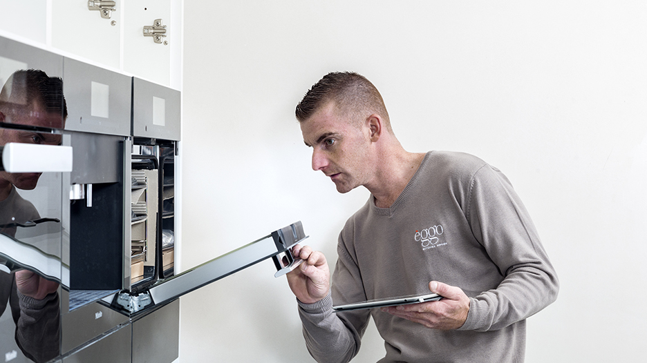Un homme en pull beige inspecte un four encastré noir avec une tablette à la main dans une cuisine moderne.
