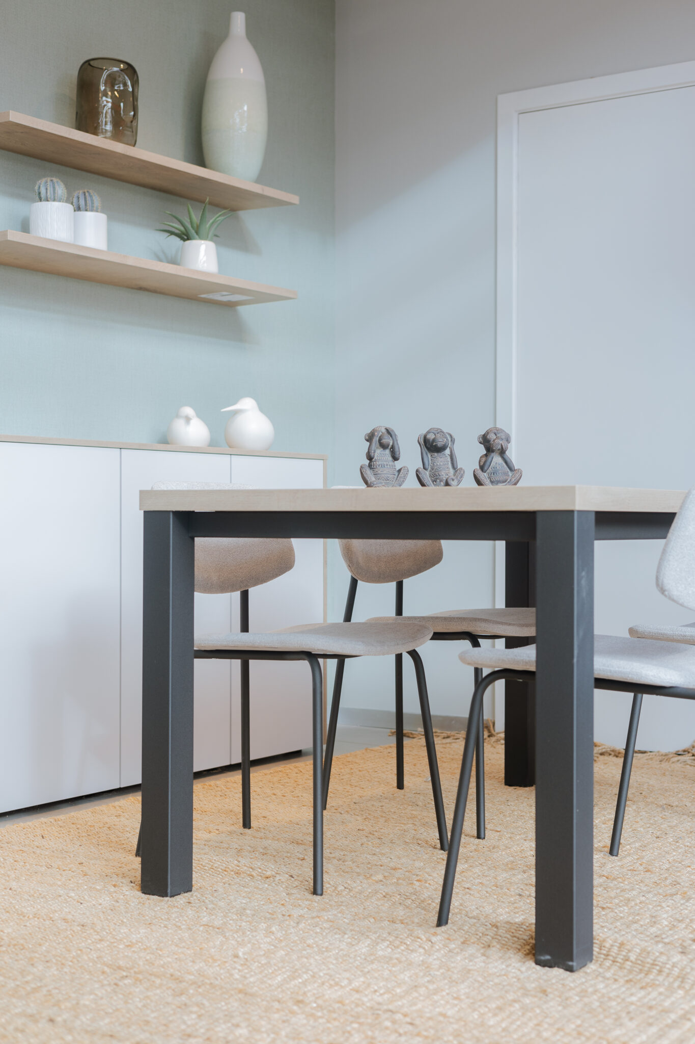 Une table en bois clair avec des chaises grises, décorée de trois statuettes, est placée sur un tapis beige à côté d'une étagère murale.