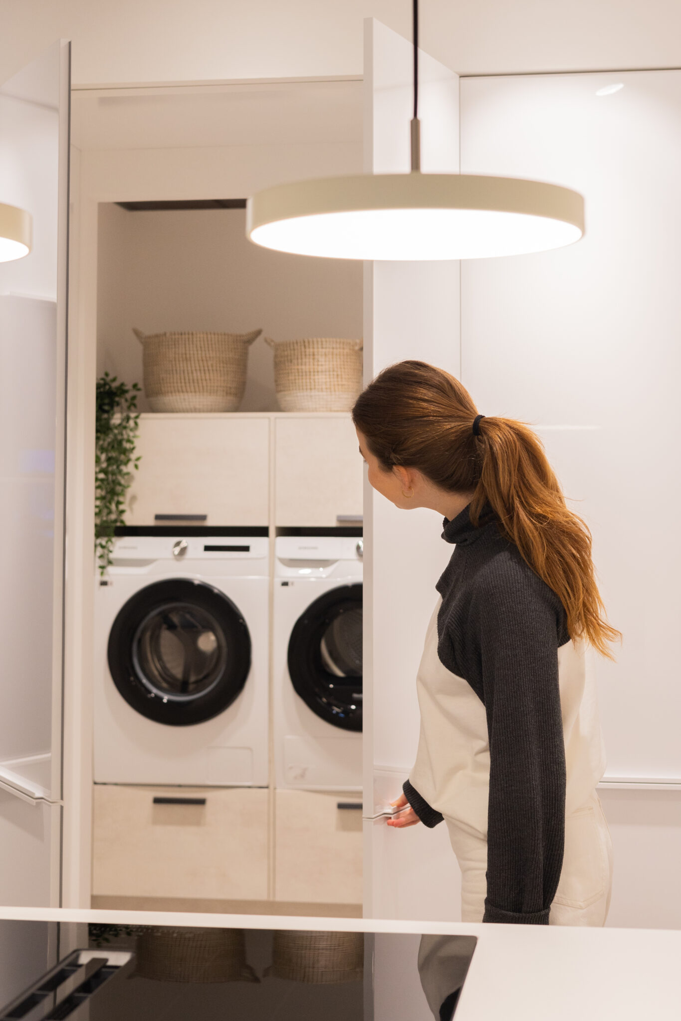 Une femme ouvre une porte vers une buanderie avec une machine à laver et un sèche-linge blancs, des paniers en osier au-dessus.