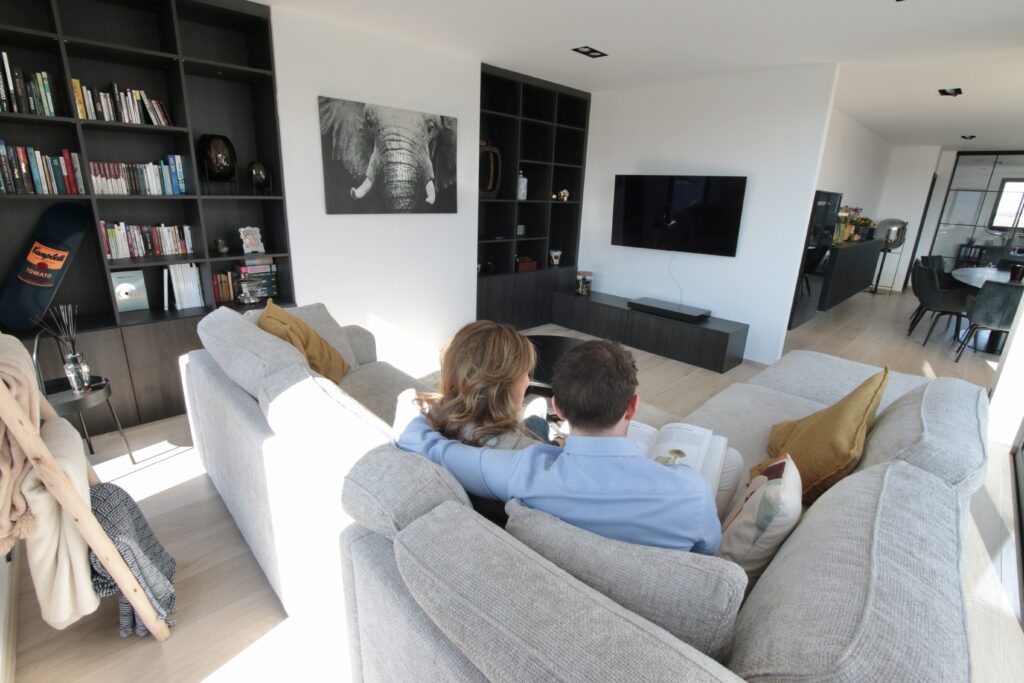 Un couple est assis sur un canapé gris clair, face à une télévision murale, avec des étagères et un tableau d'éléphant en arrière-plan.