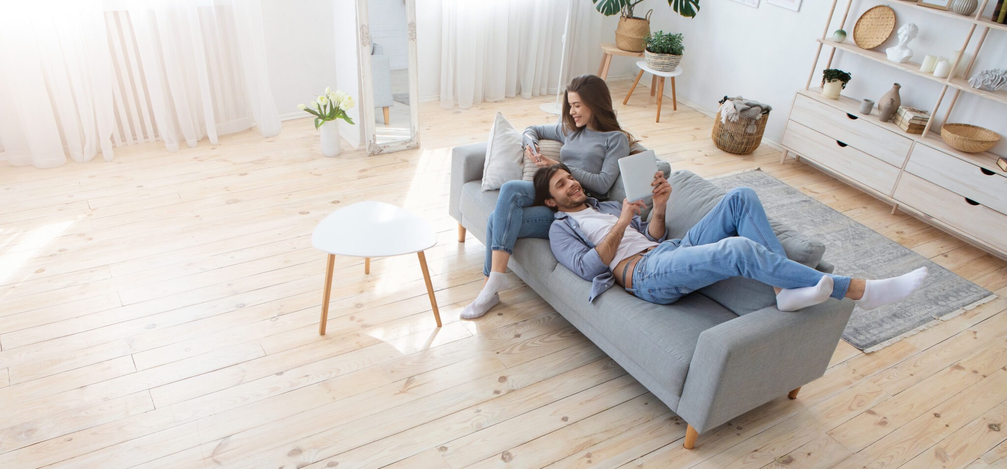 Couple détendu sur un canapé gris, lisant des tablettes, dans un salon lumineux avec sol en bois clair et décoration minimaliste.