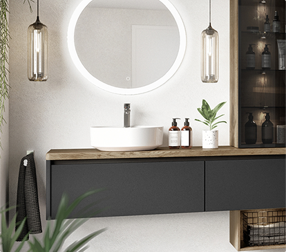 Un lavabo blanc sur un meuble noir avec un miroir rond éclairé, deux suspensions en verre et des plantes vertes.