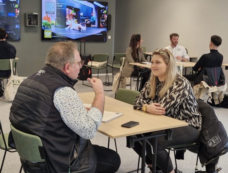 Deux personnes discutent à une table dans une salle, avec des écrans accrochés au mur en arrière-plan.