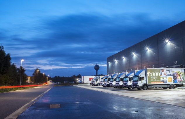 Camions alignés devant un entrepôt éclairé, sous un ciel bleu nocturne, avec des lampadaires illuminant la route à gauche.
