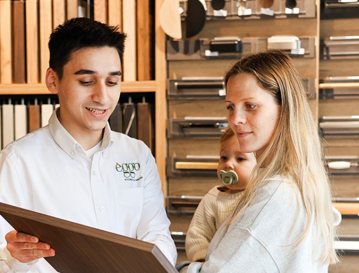 Un homme en chemise blanche montre un échantillon de bois à une femme tenant un enfant dans un showroom de cuisine.